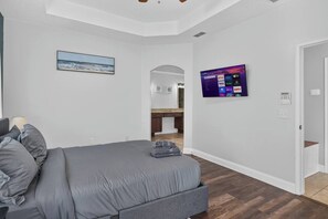 A bedroom with a bed, a TV mounted on the wall, and a view into the kitchen. - 3BR Pool Home/Close to Beaches/Mayo/UNF/Towncenter (Jacksonville)