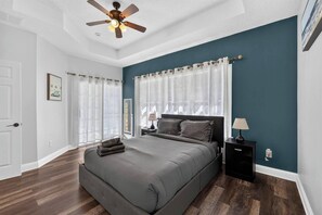 Un dormitorio con una cama, una mesita de noche, un ventilador de techo y una ventana con cortinas.