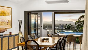 A dining area with a table and chairs, a large window with a view of a pool and mountains, and a framed artwork on the wall. - Noosa Sunsets - Luxury Home with Stunning Views (Noosa Heads)