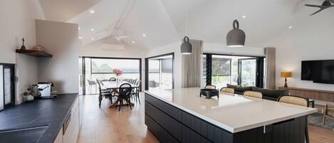 A modern kitchen with a large island, dining area with chairs, and a sofa in a living area with a TV.