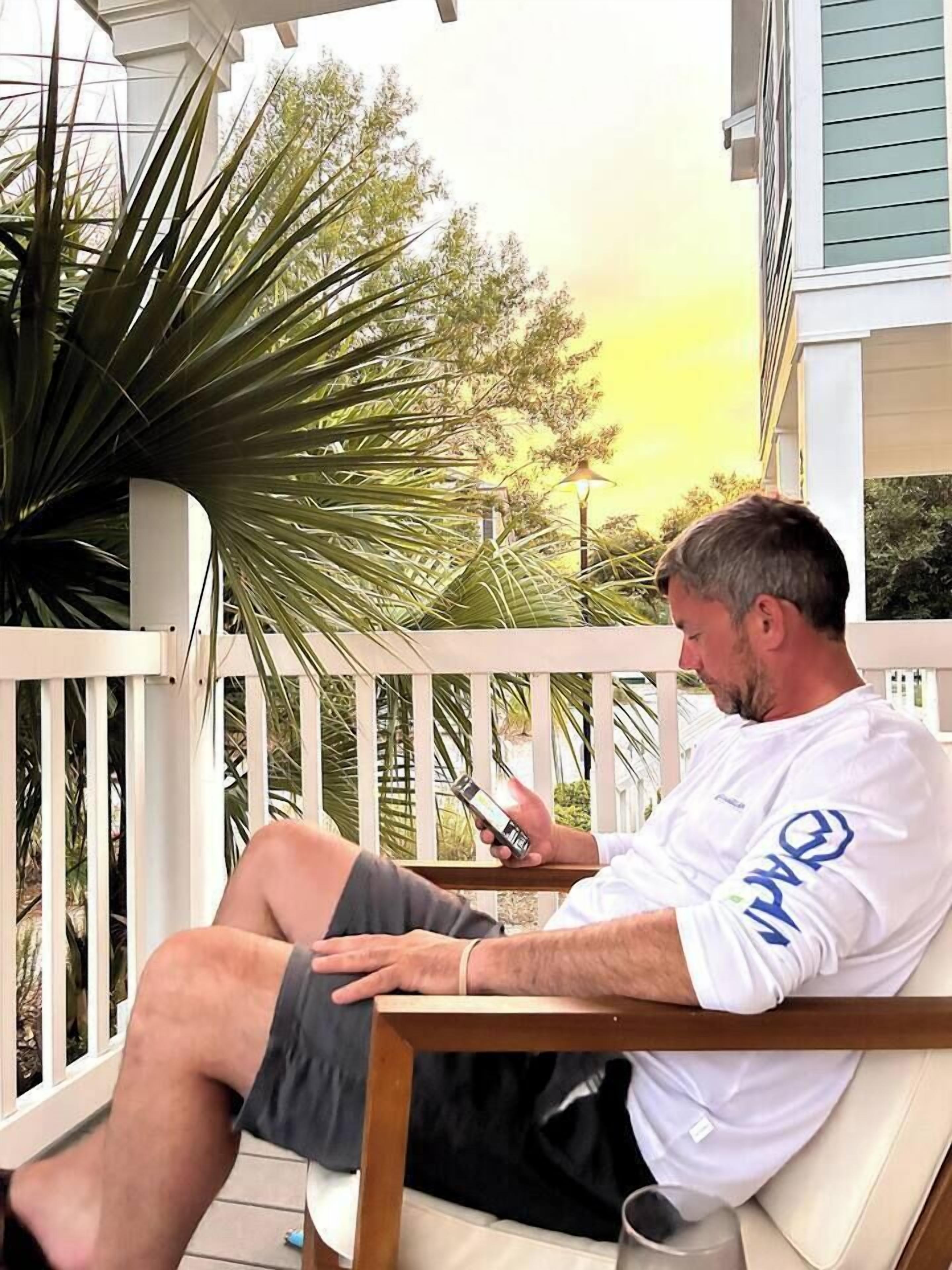A man sitting on a wooden chair on a porch with a white railing, using a smartphone.
