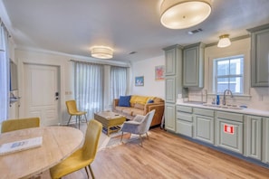 Un espace de vie compact comprenant un coin repas, un canapé, une cuisine avec des meubles de couleur vert clair et une fenêtre avec des stores.