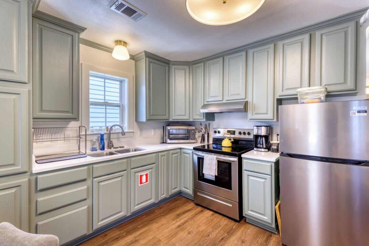 A kitchen with light green cabinets, stainless steel appliances, a window with blinds, and a wooden floor.