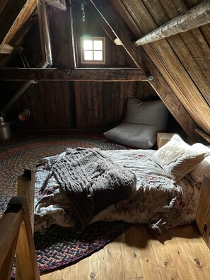 Une chambre de style loft avec un plafond en pente, une fenêtre, un lit recouvert d’une courtepointe à motifs, un oreiller gris et un plancher en bois.
