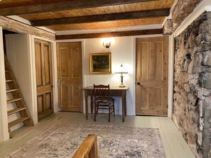 Une pièce avec un bureau en bois, une chaise et une lampe. Un escalier se trouve à gauche et un mur en pierre à droite.