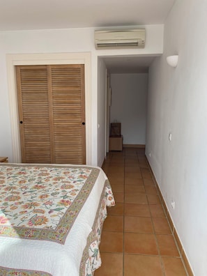 Quarto com uma cama, um guarda-roupa de madeira, ar condicionado e um corredor que leva a um banheiro.