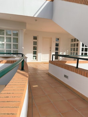 Um corredor com piso ladrilhado, uma porta branca e uma janela com vista para o céu.