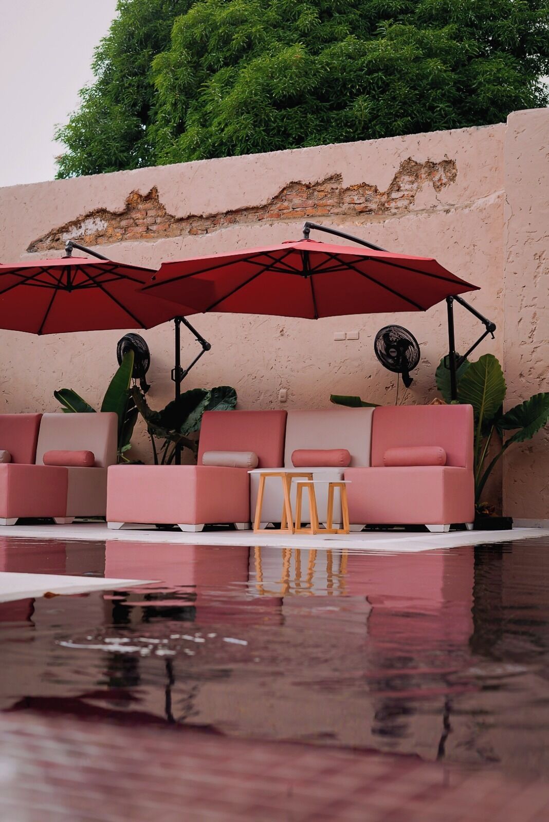 A poolside seating area with two red umbrellas, a fan, and a sofa with cushions.