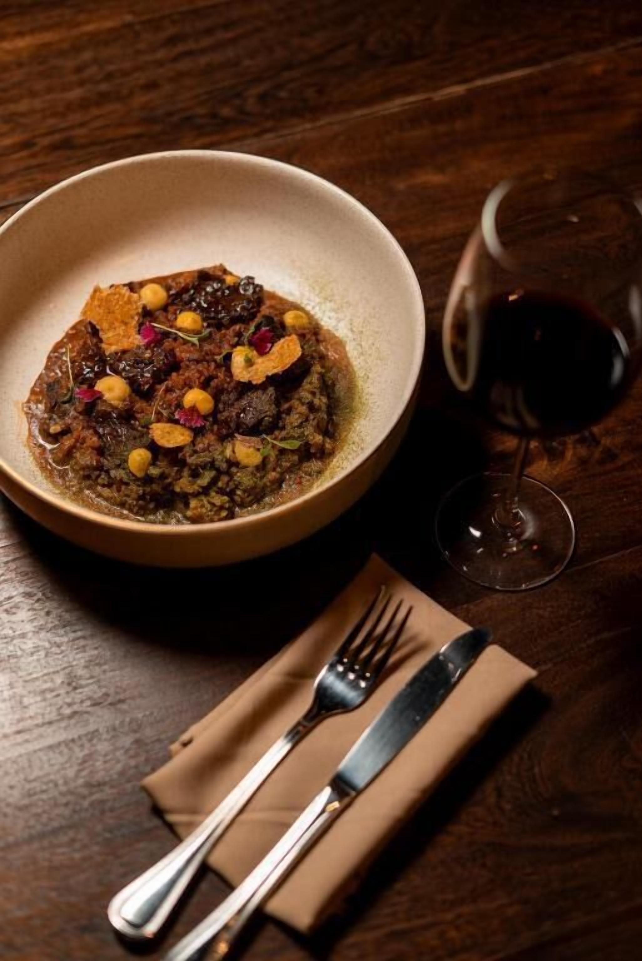 A bowl of food with a glass of red wine on a wooden table.