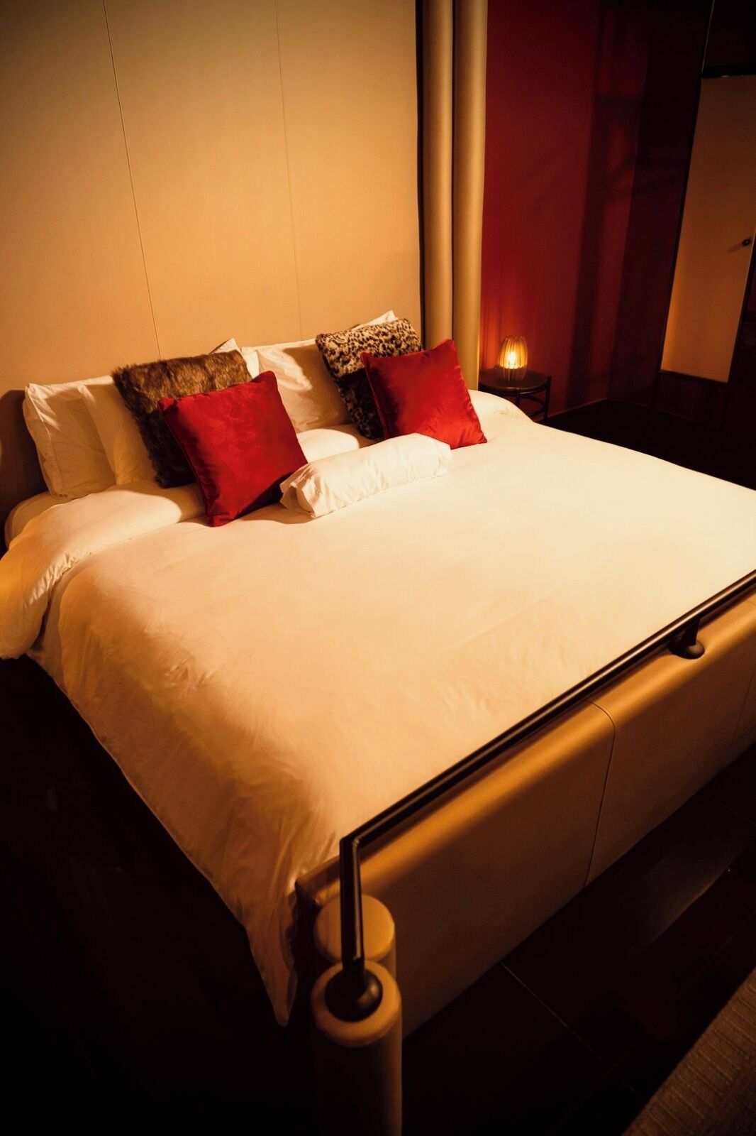 A bed with white bedding and multiple pillows, including red and patterned ones, in a dimly lit room with a nightstand and a lamp.