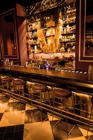 Una zona de bar con suelo a cuadros, taburetes metálicos y una estantería llena de botellas.