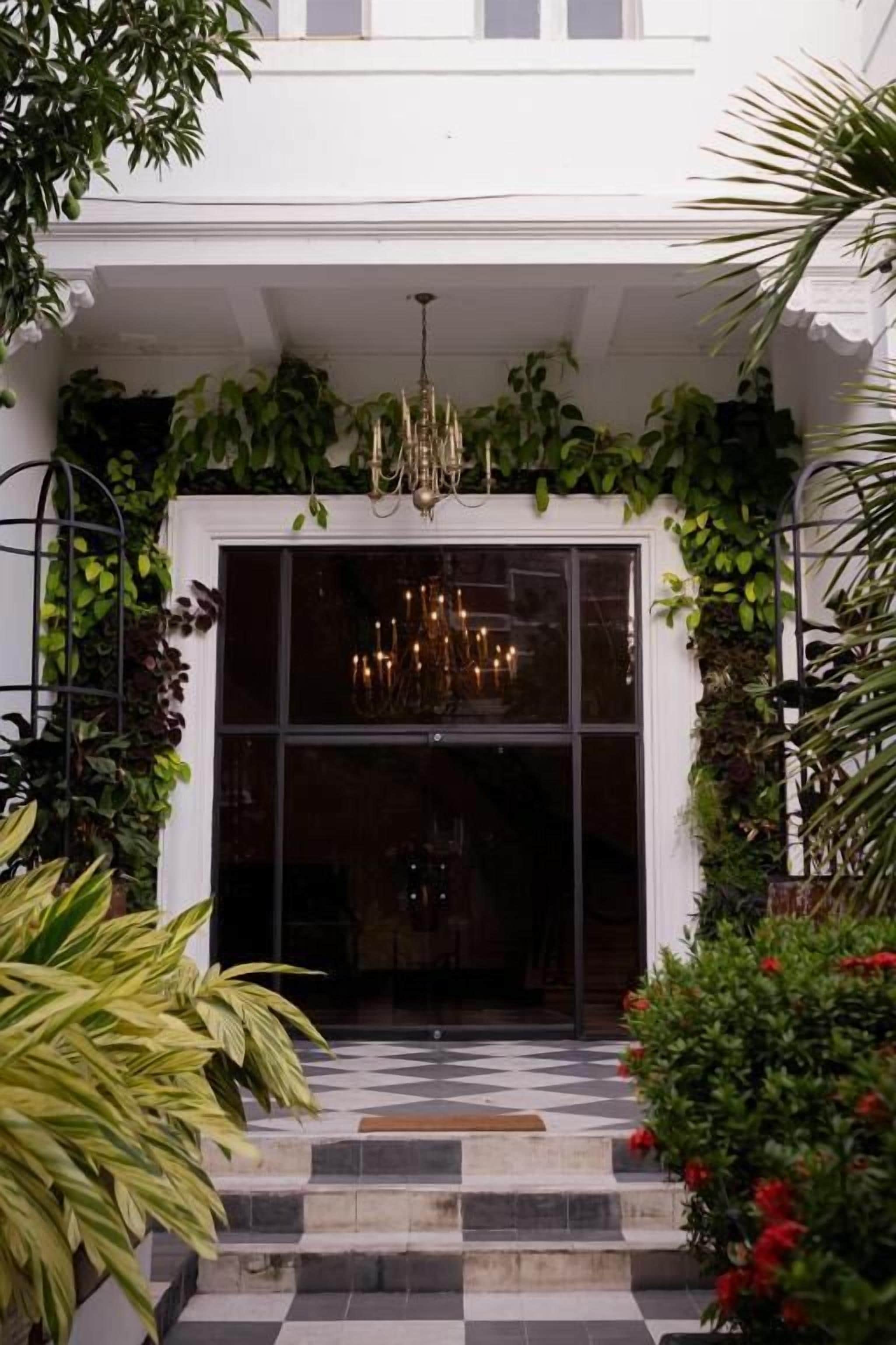 Entrada de hotel com piso xadrez, um candelabro e vegetação exuberante.