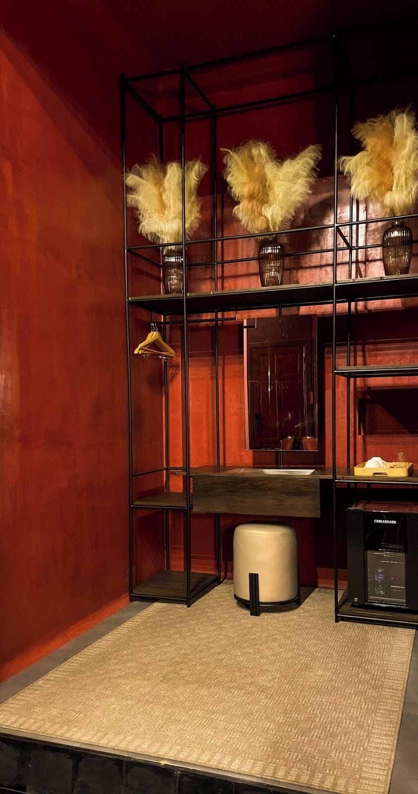Um quarto com uma estante de metal preto, um banquinho bege e um pequeno eletrodoméstico no chão.