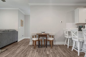 A modern living room with a grey sofa, a wooden dining table with four chairs, and a kitchen area with white cabinets and a countertop. - Brand NEW Calabash Condo (Calabash)