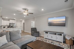 A modern living room with a grey sofa, a coffee table, a grey armchair, a flat-screen TV mounted on the wall, and a kitchen area in the background. - Brand NEW Calabash Condo (Calabash)