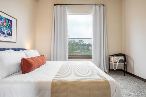 A hotel room with a bed, a window with curtains, a chair, and a wall-mounted bag rack.