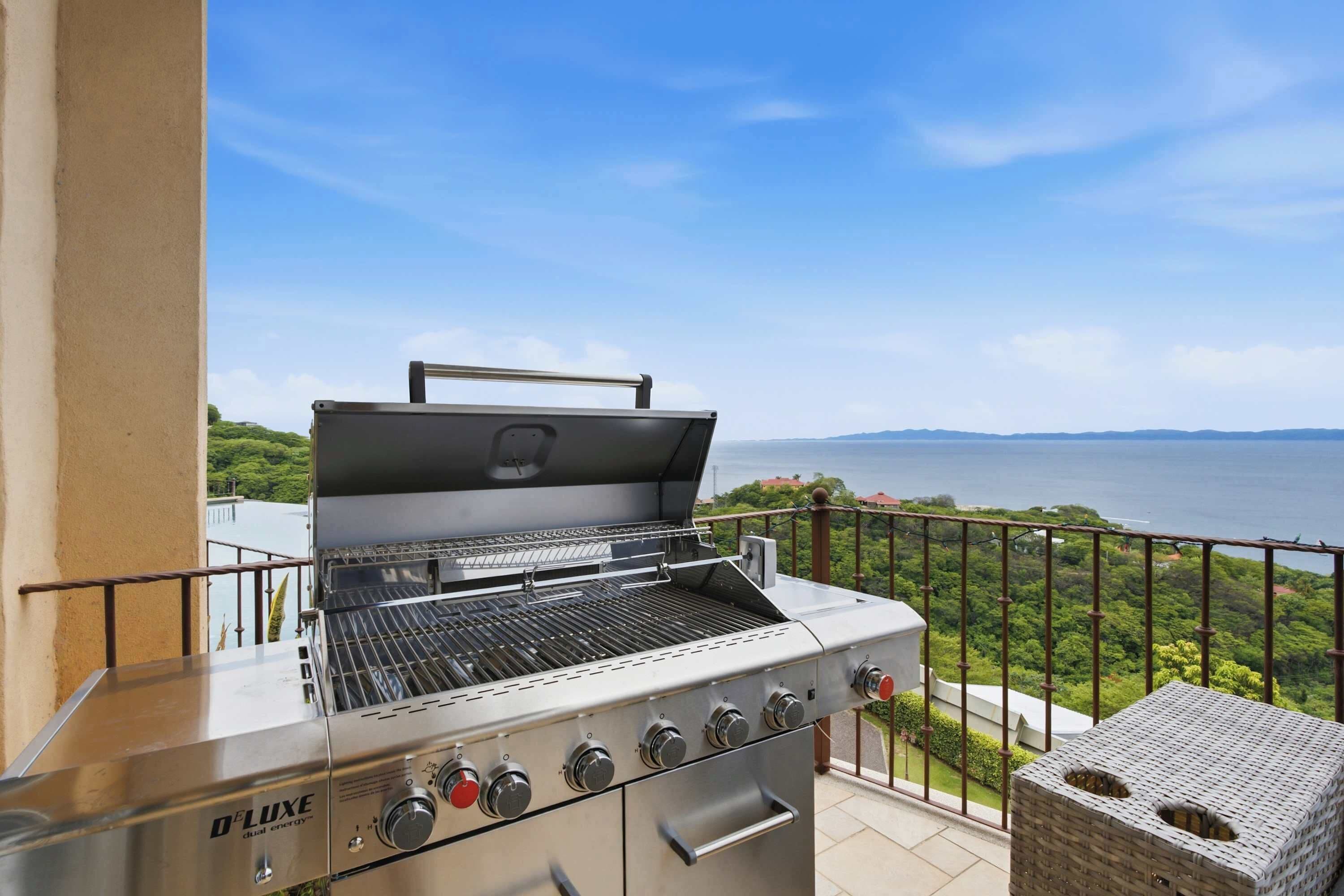 A D-Luxe dual energy grill with multiple burners and a chimney, situated on a balcony with a view of a body of water and greenery.