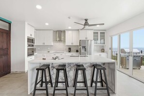 A modern kitchen with white cabinetry, a marble island with bar stools, and a view of the outdoors through large sliding glass doors. - Ocean & Mountain Views, Pool in Playa Ocotal (Playas del Coco)