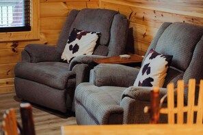 Une pièce chaleureuse avec deux fauteuils gris à revêtement en tissu et dotés de coussins en peau de vache, une petite table en bois et une fenêtre avec des stores.