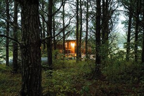 Une cabane au cœur d’une forêt, dotée d’une terrasse bien visible et d’une source de lumière chaleureuse.