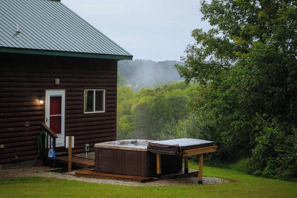 Una cabaña de madera con jacuzzi en la parte delantera, rodeada de vegetación.