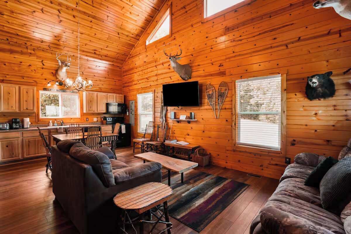 Interior de una cabaña con paredes de madera, chimenea, televisor y un corzo colgado.