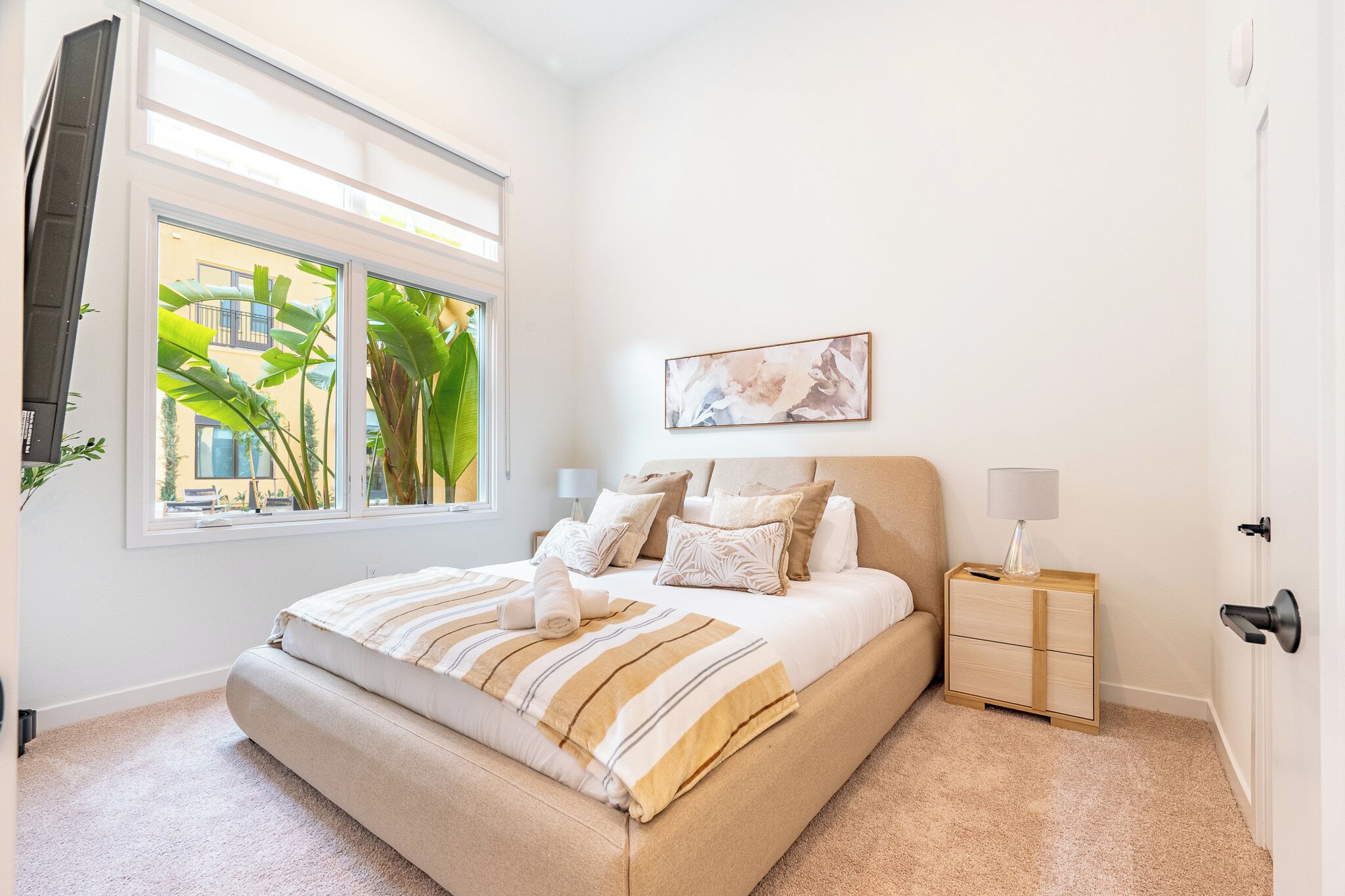 A bedroom with a large bed, a bedside table, a nightstand, a window with a view of buildings, and a painting on the wall.