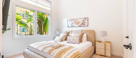 A bedroom with a large bed, a bedside table, a nightstand, a window with a view of buildings, and a painting on the wall.