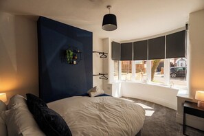 A bedroom with a large bed, a dark accent wall, a window with blinds, and a bedside table with a lamp. - Cozy 4BR Redcar House | Sleeps 7 | Close to Beach (Redcar and Cleveland)