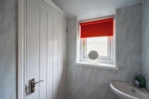 A bathroom with a white door, a window with a red blind, a round mirror, and a sink. - Cozy Blue Ember Stay | Work friendly 4BR+Sleeps 7 (Redcar and Cleveland)