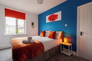 A bedroom with a bed, a bedside table with a lamp, a window with a red blind, and a wall with a large flower painting. - Cozy Blue Ember Stay | Work friendly 4BR+Sleeps 7 (Redcar and Cleveland)
