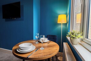 A dining area with a round wooden table set for two, a television mounted on the wall, a floor lamp, and a window with a plant on the sill. - Cozy Blue Ember Stay | Work friendly 4BR+Sleeps 7 (Redcar and Cleveland)