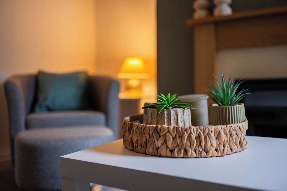 A modern living room with a white table, a woven tray with plants, a sofa, and a lamp.