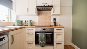 A kitchen with white cabinets, a stainless steel oven, a green kettle, and a window with a plant. - The Alfred Suite | Redcar (Redcar and Cleveland)