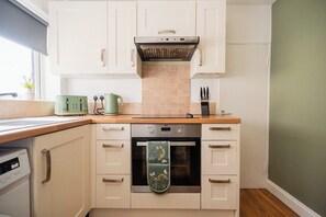 A kitchen with white cabinets, a stainless steel oven, a green kettle, and a window with a plant. - The Alfred Suite | Redcar (Redcar and Cleveland)