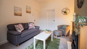 A living room with a grey sofa, a white coffee table, and a grey armchair. There are two doors, a wall clock, and a plant on a wooden stand. - The Alfred Suite | Redcar (Redcar and Cleveland)