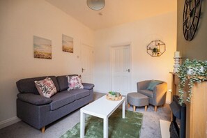 A living room with a grey sofa, a white coffee table, and a grey armchair. There are two doors, a wall clock, and a plant on a wooden stand. - The Alfred Suite | Redcar (Redcar and Cleveland)