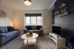 A modern living room with two sofas, a coffee table, a television on a stand, and a window with a view of buildings. - 3BR Barnaby House | Contractor Friendly | Sleeps 5 (Redcar and Cleveland)