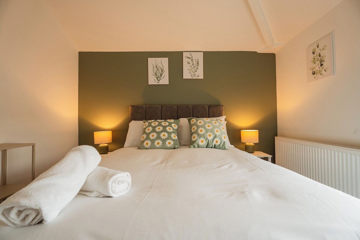 A neatly made bed with a white duvet, two pillows with a daisy pattern, and a rolled white towel on the side. There are two bedside lamps on either side of the bed.