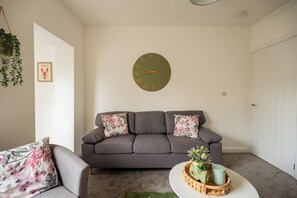 Ein Wohnzimmer mit grauem Sofa, floralen Kissen, einer runden Wanduhr und einem kleinen Tisch mit Pflanze und Kerze.