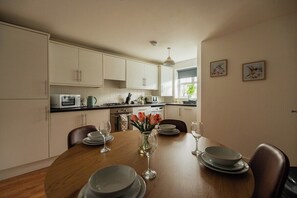 A modern kitchen with white cabinets, a wooden dining table set for four, and a vase with flowers. - 3BR Einstein House in Hardwick | Work Friendly (Stockton-on-Tees)