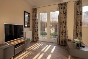 Ein modernes Wohnzimmer mit einem Flachbildfernseher, einer Holz-Unterhaltungsecke, einer Schiebetür zum Garten und einem kleinen Tisch mit einer Pflanze.
