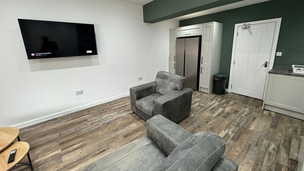 A living room with a flat-screen TV mounted on the wall, grey sofas, a wooden floor, and a kitchen area with cabinets and a door. - 6 Bed Contractor Acom in Hartlepool (Hartlepool)