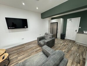 A living room with a flat-screen TV mounted on the wall, grey sofas, a wooden floor, and a kitchen area with cabinets and a door. - Modern 6 Bed Stay in Hartlepool | Work friendly (Hartlepool)