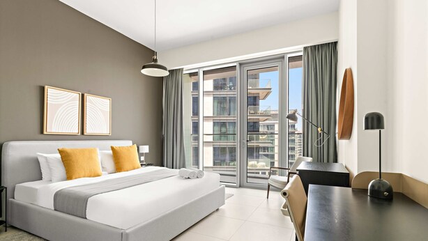 A modern hotel room with a bed, a desk, a chair, and a view of the city through large windows. - Silkhaus Premium 2bdr Stay in Canal Front (Dubai)