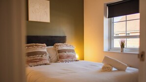 Una cama bien hecha con almohadas y una toalla enrollada, una ventana con vistas a edificios y un cuadro en la pared.