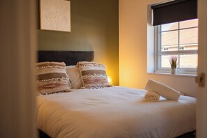 A neatly made bed with pillows and a rolled towel, a window with a view of buildings, and a framed picture on the wall. - The Norton Nook | Horizon Stays (Stockton-on-Tees)