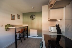 A compact kitchen with a wooden countertop, a wall-mounted clock, a radiator, and a small dining area with stools. - 3BR Norton Nook | Sleeps 5 | Work Friendly + WiFi (Stockton-on-Tees)