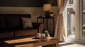 Un salón con un sofá marrón oscuro, una mesa de centro de madera con una bandeja de objetos, una lámpara en una mesita auxiliar y vistas a través de una ventana con cortinas.