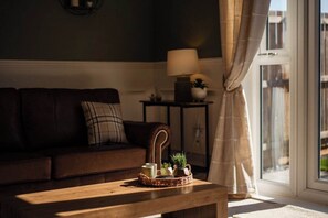 A living room with a dark brown sofa, a wooden coffee table with a tray of items, a lamp on a side table, and a view through a window with curtains. - The Norton Nook | Horizon Stays (Stockton-on-Tees)